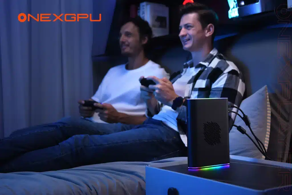 Two men sitting on a couch gaming with controllers in hand, illuminated by ambient blue lighting. A sleek external GPU device, the ONEXGPU 2, sits on a nearby table, featuring RGB lighting and multiple connected cables. The ONEXGPU logo is prominently displayed.