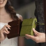 Two individuals holding compact leather bags One bag is green and the other is brown both featuring the AYANEO logo and a sleek design The setting suggests casual or professional usage