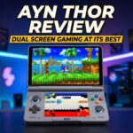 The image features a dual screen clamshell style handheld gaming device centered on a dark surface against a blurred neon lit gaming room background The device is grey with colorful ABXY buttons and glowing blue rings around the joysticks The top screen displays a vibrant 3D Sonic the Hedgehog game while the bottom screen shows a stylized menu or secondary game interfaceThe image features a dual screen clamshell style handheld gaming device centered on a dark surface against a blurred neon lit gaming room background The device is grey with colorful ABXY buttons and glowing blue rings around the joysticks The top screen displays a vibrant 3D Sonic the Hedgehog game while the bottom screen shows a stylized menu or secondary game interface