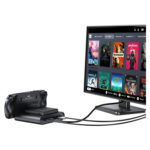 A black GPD WIN 5 handheld console is shown docked and connected via cables to a monitor The monitor displays the Steam OS style interface with a grid of game titles such as Dead Cells Deathloop Portal 2 and Stardew Valley illustrating the devices ability to drive a desktop gaming experience