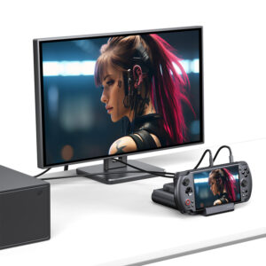 A desk setup showing the GPD WIN 5 docked and connected to an external monitor via cables. The same image of a female character with pink hair is displayed on both the small console screen and the large external monitor, demonstrating the dock's video output capability.