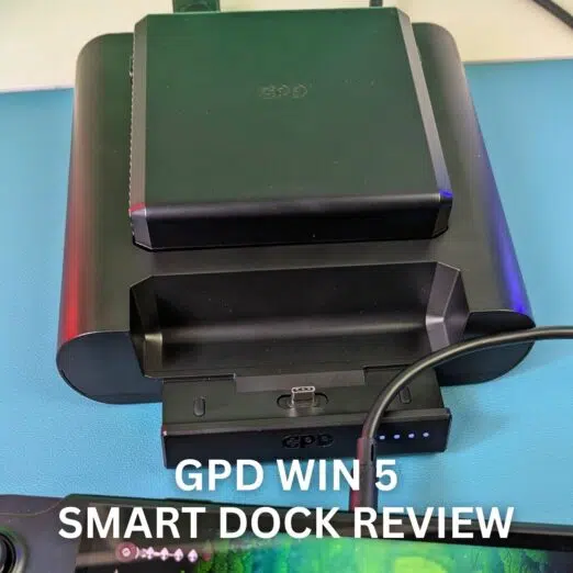 GPD WIN 5 SMART DOCK REVIEW 11 A close-up photograph of the black GPD WIN 5 Smart Dock sitting on a light blue surface. The dock is designed to hold the GPD WIN 5, with the console's screen partially visible in the foreground, displaying a gaming interface. The dock itself is connected by a black cable, and its LED indicator lights are visible near the charging port. Large white text overlaid on the image reads, "GPD WIN 5 SMART DOCK REVIEW."