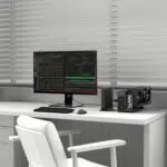 Desk setup with a widescreen monitor displaying code and terminal windows, a compact PC and external GPU enclosure on the right, white chair and blinds in the background