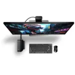 GPD BOX mini PC mounted beneath an ultrawide monitor, black ribbed case with keyboard, mouse, and webcam