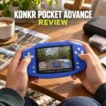 A person holding a blue handheld gaming device styled like a Game Boy Advance, playing a 3D rally racing game. Large bold text at the top reads "KONKR POCKET ADVANCE" in white with "REVIEW" highlighted in lime green below. The device is displayed on a wooden desk alongside a cup of coffee, game cartridges, and a notebook in a cozy indoor setting.
